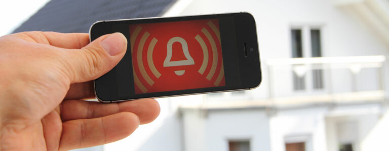 Ein Smartphone zeigt einen Einbruchalarm in einem dahinter stehenden Haus an.